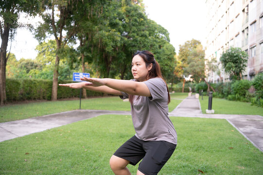 A person performs a forward-extended squat in a sunlit green park surrounded by trees and pathways.