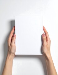 Hands holding a blank, white rectangle against a stark white surface, softly lit from above and beside