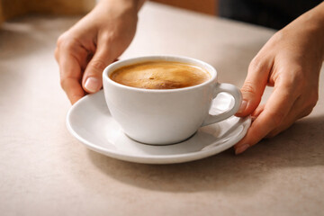 A person's hands holding a white cup of freshly brewed coffee with crema on a saucer on a light table.