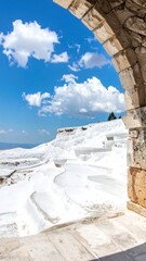 A stone arch frames a picturesque view of layered, white mineral formations under a vibrant blue sky dotted with puffy clouds