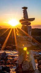 A stone cairn stands silhouetted against a brilliant, radiant sunset, casting dramatic light rays across the rugged, mountainous landscape
