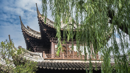 A traditional old Chinese building against a blue sky and clouds. Tiled roof with curved cornice edges. Latticed terrace railings. Green willow branches in the foreground. China. Shanghai. © Вера 