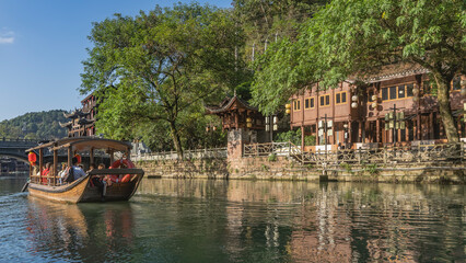 Naklejka premium A tourist wooden boat is sailing along a picturesque river. There are traditional Chinese buildings and green vegetation on the shore. The fence along the embankment. Reflection on the water. China.