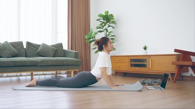 Young Asian woman stretching yoga workout on exercise mat while online training class with computer laptop in living room