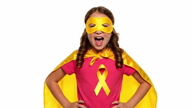 A young girl wearing a pink t-shirt with a yellow ribbon, yellow cape, and matching mask, posing heroically on International Childhood Cancer Day for awareness of sarcoma in February.
