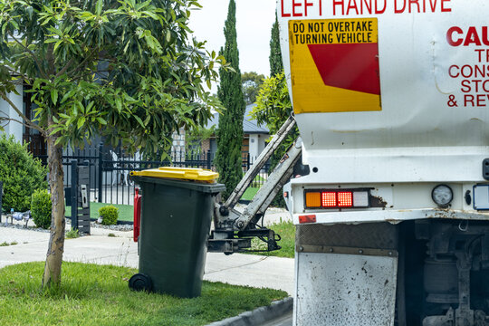 A municipal garbage truck collecting a household wheelie bin along a quiet suburban street in Australia. Routine waste collection, local council services, and residential infrastructure