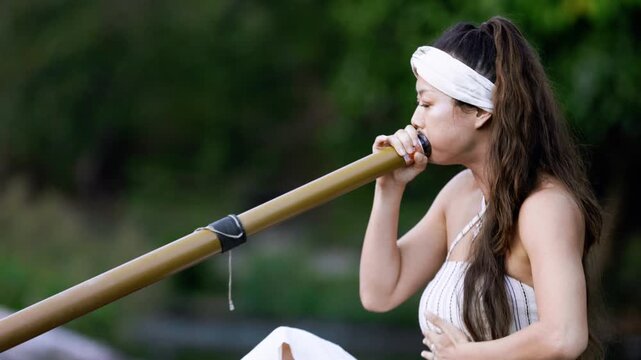 A peaceful moment captured in nature as a woman skillfully plays a traditional wind Instrument didgeridoo, connecting with the surroundings and expressing her artistry and emotions.