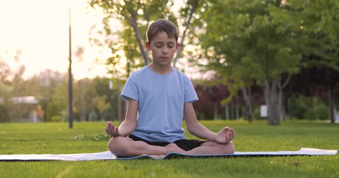 Calm child sits peacefully on green grass in park at sunset. Child boy enjoys quiet moment outdoors showing emotional balance and focus. Child wellbeing concept, healthy childhood and mental stability