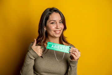 Happy woman smiling, holding an abierto (open) sign, and giving a thumbs up gesture, indicating business is open