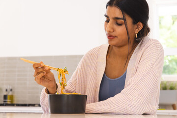 Asian woman sitting at kitchen counter using chopsticks lifting noodles from black bowl, copy space