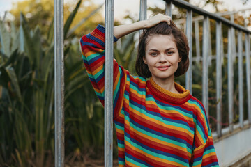 Fototapeta premium Young woman in her twenties wearing a colorful striped sweater, smiling and leaning on a metal railing in an urban garden with short hair, casual outfit and relaxed lifestyle portrait.