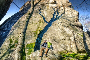 Hiker resting by giant rocks. Man with backpack under towering ancient cliff	
