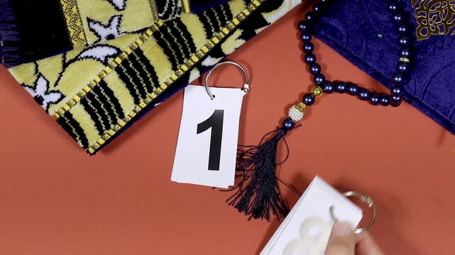 Close Up Calendar Tags With February 18 which mean ramadan day with prayer mat and A closed copy of the Holy Quran with Arabic calligraphy on a blue cover. The Arabic text reads Al-Qur&rsquo;an Al-Kareem wh