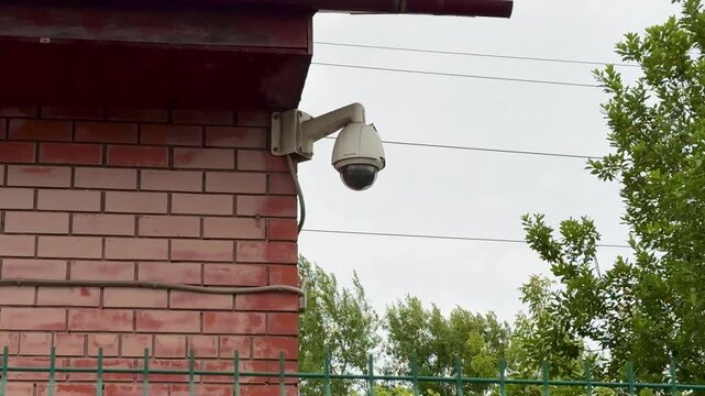 Outdoor Dome Camera Mounted On Brick Overlooking Fenced Residential Perimeter And Street, Wired To Power Lines With Green Trees And Cloudy Sky In Background, Facility Manager Monitoring