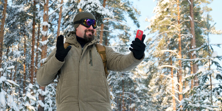 Man beaming in a winter forest, wearing ski goggles and a jacket, holding a smartphone for a video call or selfie, sharing his snowy adventure and connecting during outdoor leisure