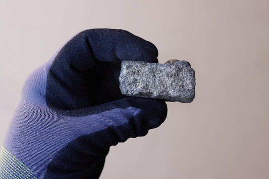 Hand with safety glove holding pentlandite ore against tan background. Source of Nickel (Ni) metal. Critical mineral, raw material. Used in battery cathodes. Mine, mining