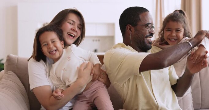 Happy interracial parents tickling their two laughing biracial children playing together on couch in living room, spending family time at home, showing unity and warmth. Leisure, playtime with kids
