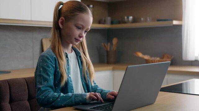 Caucasian little school girl child kid daughter at home kitchen using family laptop chatting website playing gaming using internet browsing children and modern technology generation studying learning