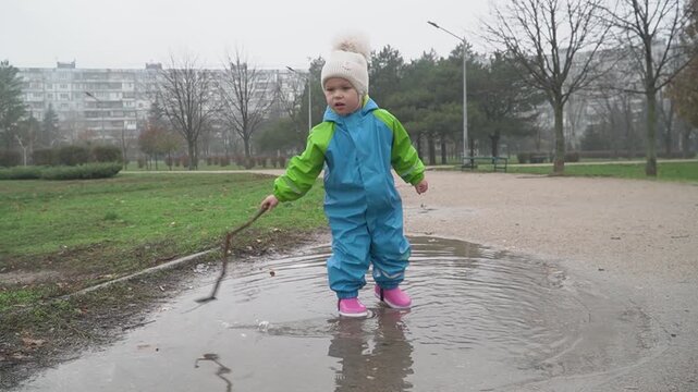 A girl in a blue and green jumpsuit and pink rubber boots in a white warm hat hits a large puddle in the park with a long stick, stands in it, an autumn rainy day, cold and damp, looks around