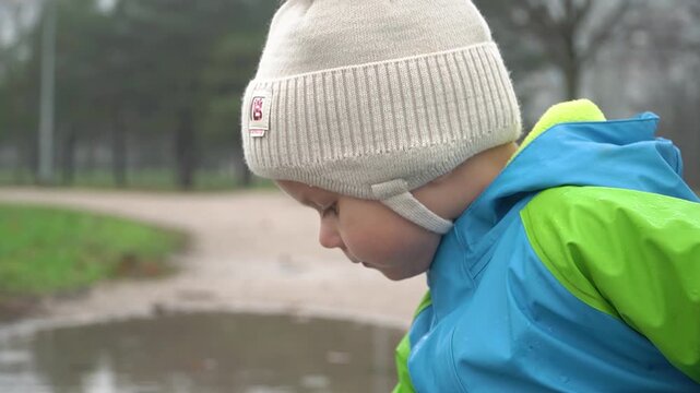A child in a white warm hat and a blue raincoat plays in a park near a puddle in the autumn rain, focused and attentive, a gray day, cold