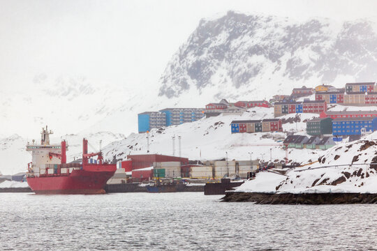 Arctic harbor scene with a cargo ship docked at the waterfront, colorful Inuit houses on a snowy hillside with mountains in background, Sisimiut, Greenland
