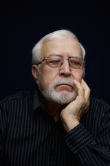 Obraz premium Older man with glasses holding his chin while deep in thought in a dark setting