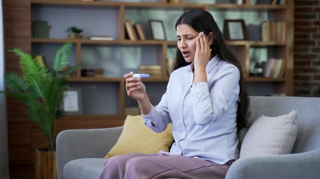 Stressed young woman feeling unhappy after looking on positive pregnancy test results while sitting on sofa in living room at home. Female being worried about unplanned pregnancy, fertility problems.