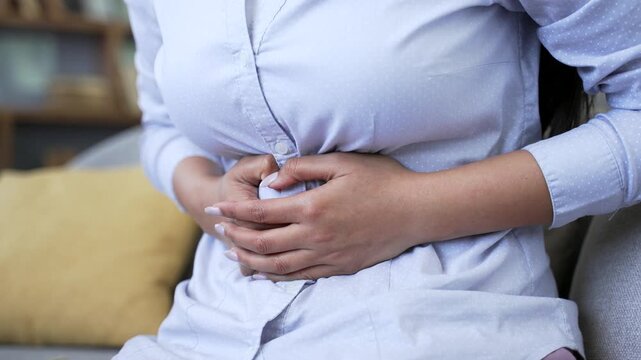 Close up woman suffering from abdominal pain sitting on sofa in living room at home. Unhealthy adult female keeps her hands on her stomach. She has poisoning, constipation, bloating or menstruation