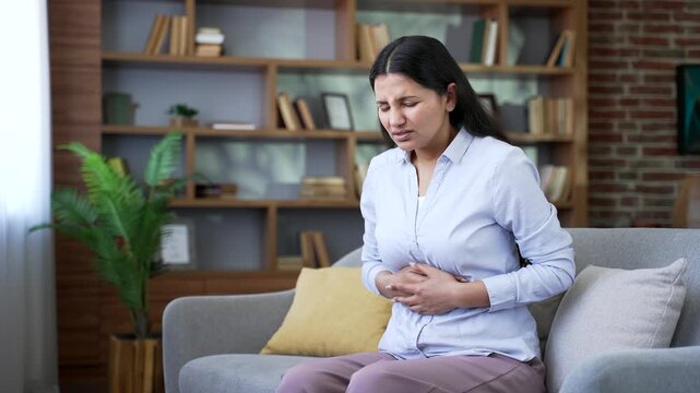 Woman suffering from abdominal pain sitting on sofa in living room at home. Unhealthy female keeps her hands on her stomach. Girl has poisoning, constipation, bloating or menstruation.