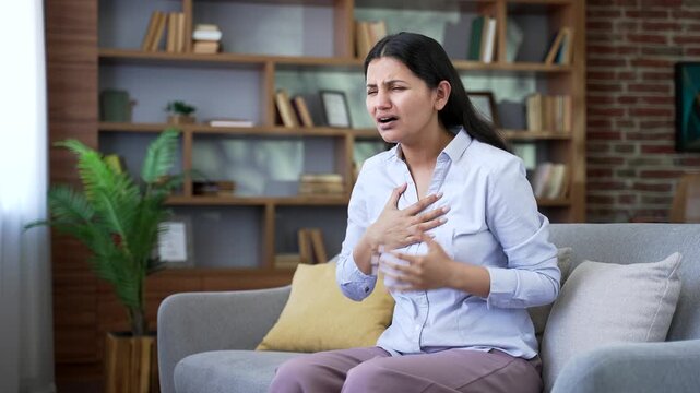 Adult woman has heart attack while sitting on sofa in living room at home. Sick young female suffers from tension in chest and massages sore spot with her hand. She needs urgent medical attention
