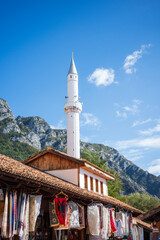 Obraz premium A vibrant outdoor souvenir market featuring colorful handmade textiles, rugs, and crafts displayed on a rustic wooden building, with a tall white mosque minaret rising against dramatic rocky mountains