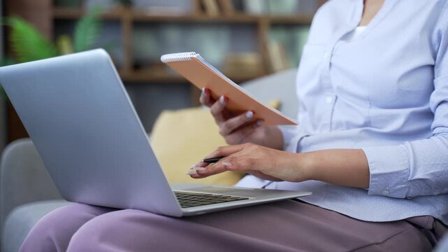 Close up woman using laptop and taking notes in notebook while sitting on the sofa in the living room at home. Female student writes during online training, listening to course or webinar, studies.