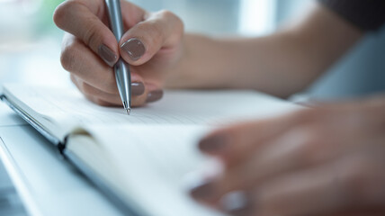 Close up of business woman hand with a pen writing on paper notebook with laptop on office desk....