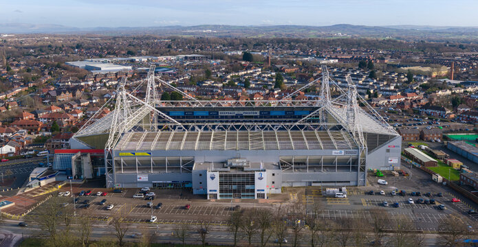 A wide aerial perspective of Preston North End's Deepdale Stadium adjacent to the historic Moor Park, showcasing the stadium's unique floodlight towers and the park's open recreational fields.