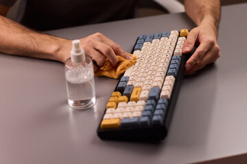 Fototapeta premium Hands cleaning mechanical keyboard with cloth, spray bottle on gray desk, closeup of keys, routine workstation hygiene, dust removal and disinfection, focused tidy atmosphere