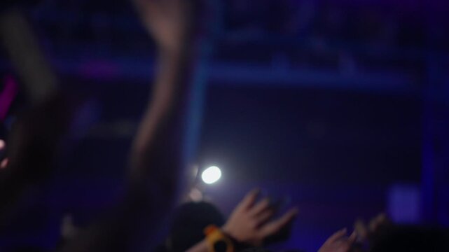 Soft-focus footage of a crowd at a live event. Fans clap and wave hands in the air under blue stage lights. A phone flashlight illuminates the scene. Energetic, emotional, and atmospheric vibe.