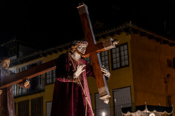 Obraz premium Jesus christ statue wearing a crown of thorns and purple robe, carrying a wooden cross, accompanied by another saint statue, during a holy week procession at night