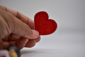 Fototapeta premium A close-up shot showing a hand holding a small, hand-drawn red heart against a neutral, light gray background.