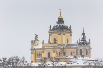 Obraz premium St George Cathedral baroque architecture view during heavy winter snowstorm. Beautiful landmark facade with golden dome under falling snow in Lviv old town. Cold cloudy evening scenic landscape.