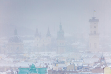 Obraz premium Panoramic view of Lviv old town landmarks under heavy snowstorm. City Hall tower, Korniakt tower and Dominican Church dome visible through blizzard. Winter urban landscape of historical center.
