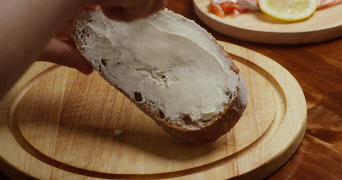 Female hands spread soft cream cheese on a slice of bread using a knife. Close-up of simple homemade breakfast preparation on a wooden board, natural light, everyday food and kitchen routine.