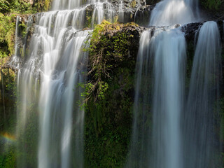 Fototapeta premium Close-up view of layered waterfall curtains flowing over mossy rock ledges in soft sunlight.