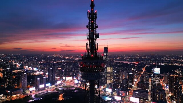 A cityscape at dusk with a tall tower in the center, surrounded by numerous buildings and streets with vibrant lights, set against a backdrop of a colorful sky with hues of blue, pink, and orange