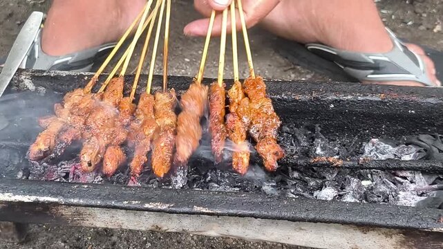 Person grilling satay or Indonesian skewers of meat on a charcoal grill in with a casual outdoor setting.