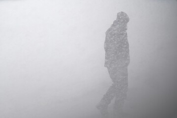 A lone figure walks through a severe blizzard, partially obscured by heavy, wind-driven snow in...