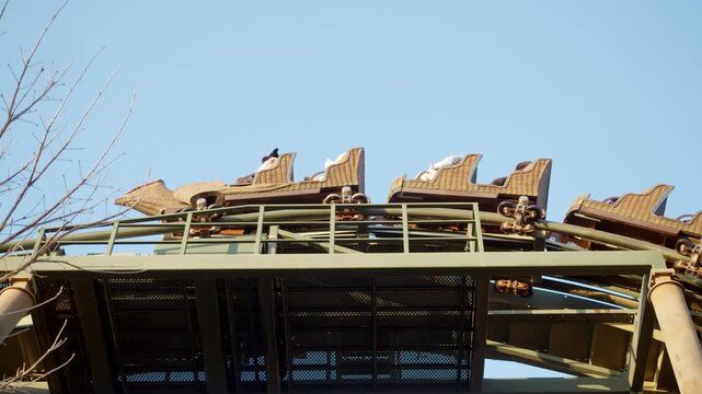 Low angle wide shot shows roller coaster train climbing curved track section above metal catwalk, with empty seats and safety restraints visible against clear blue sky.
