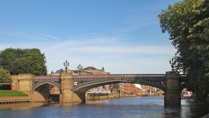 Obraz premium Historic bridge over the River Ouse in York, England, on a clear summer day. Stone arches, ornate ironwork, riverside buildings, and calm water under blue sky.