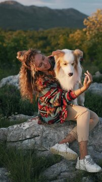 A female tourist hiker and her adorable border collie dog sit on a wild rocky terrain against a backdrop of picturesque mountain scenery. Travel, recreation, and tourism with animals, outdoor