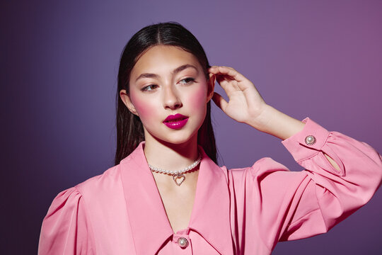 Young woman in a pink trendy outfit with bright makeup poses thoughtfully against a purple background. Concept for fashion, beauty, and style innovation.