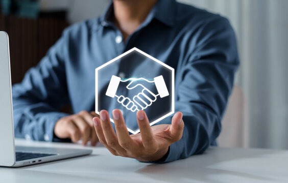 Businessman holding a glowing virtual handshake icon over an office desk. Concept of closing a deal, successful partnership, b2b agreement, trust, and professional cooperation.